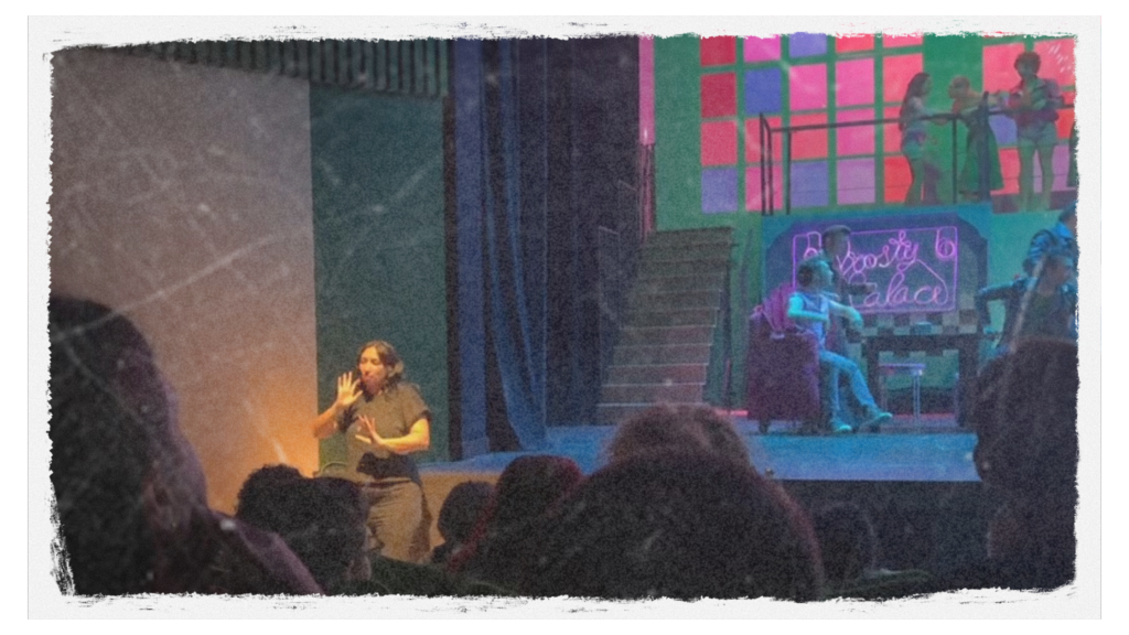 Intérprete de Lengua de Signos Española (LSE) en el extremo izquierdo de un escenario iluminado durante la representación del musical Grease en la Reunión de FAPAS CyL en Burgos. Al fondo se observa el decorado colorido y el elenco de jóvenes actuando ante el público.