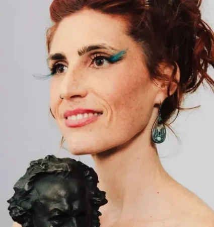 Primer plano de la actriz Miriam Garlo sonriendo de perfil mientras sostiene un premio Goya de bronce sobre un fondo gris neutro.